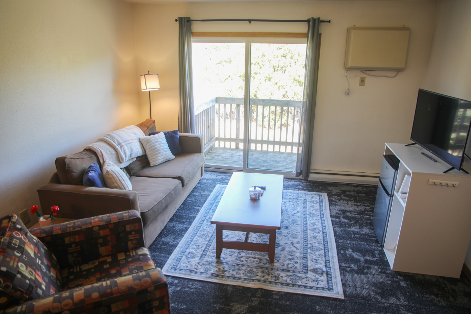 Living room with brown couches with pillows and blankets, a coffee table, with a rug and a TV stand with a mini fridge.
