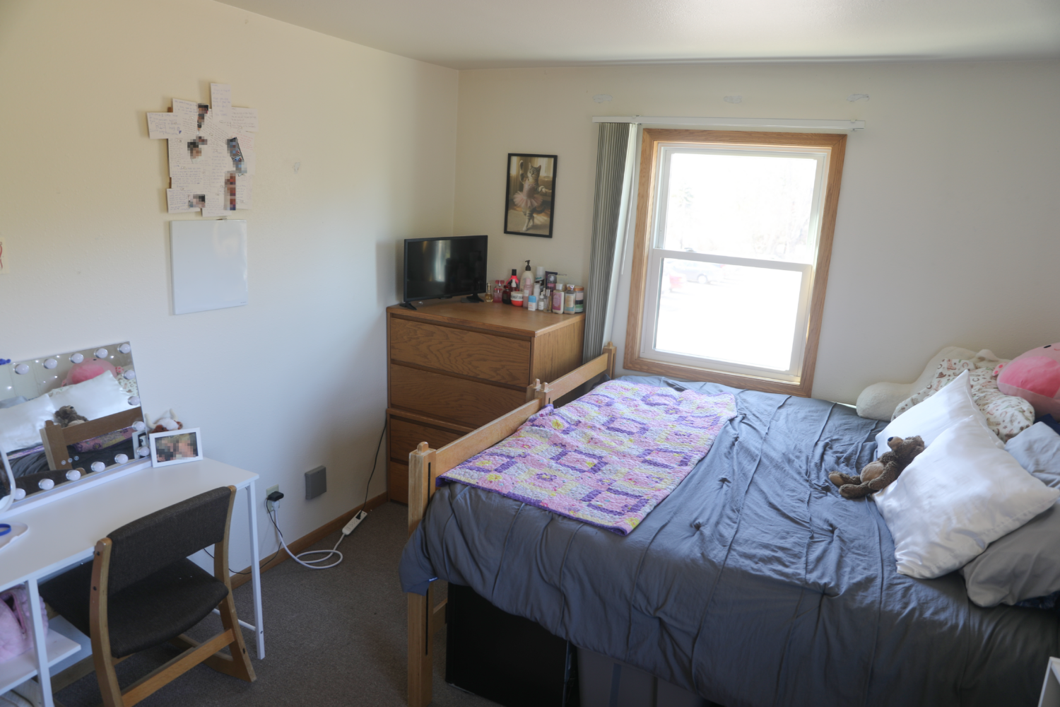 Mega bed with dark blue sheets, purple blanket, white pillows, dresser with a TV, lotions and perfumes, and a desk with a vanity.