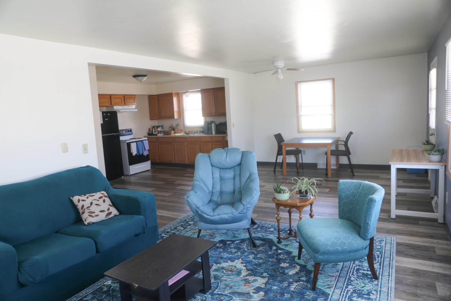 Living room and kitchen in Duplex. Blue couches, and chairs and carpet. Small dining table with 2 chairs.