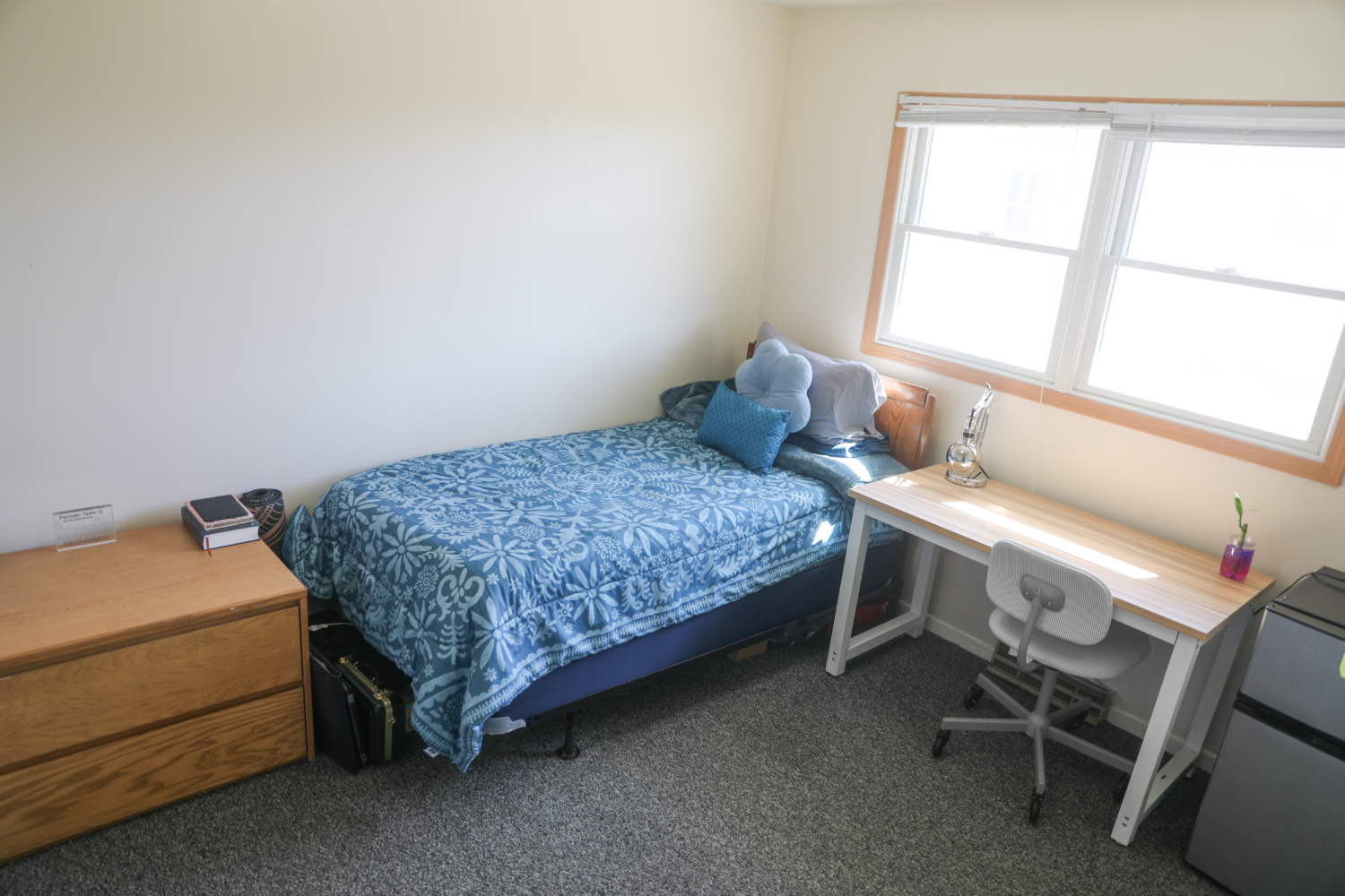 Room with a bed with blue bedding, some books on a dresser, a desk, and a mini fridge.